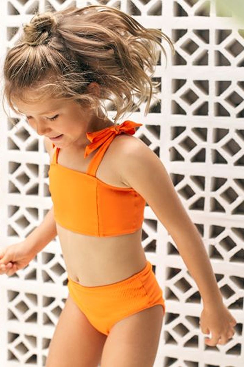 Girl in an orange bikini with a bow tie strap in front of a white breeze block wall